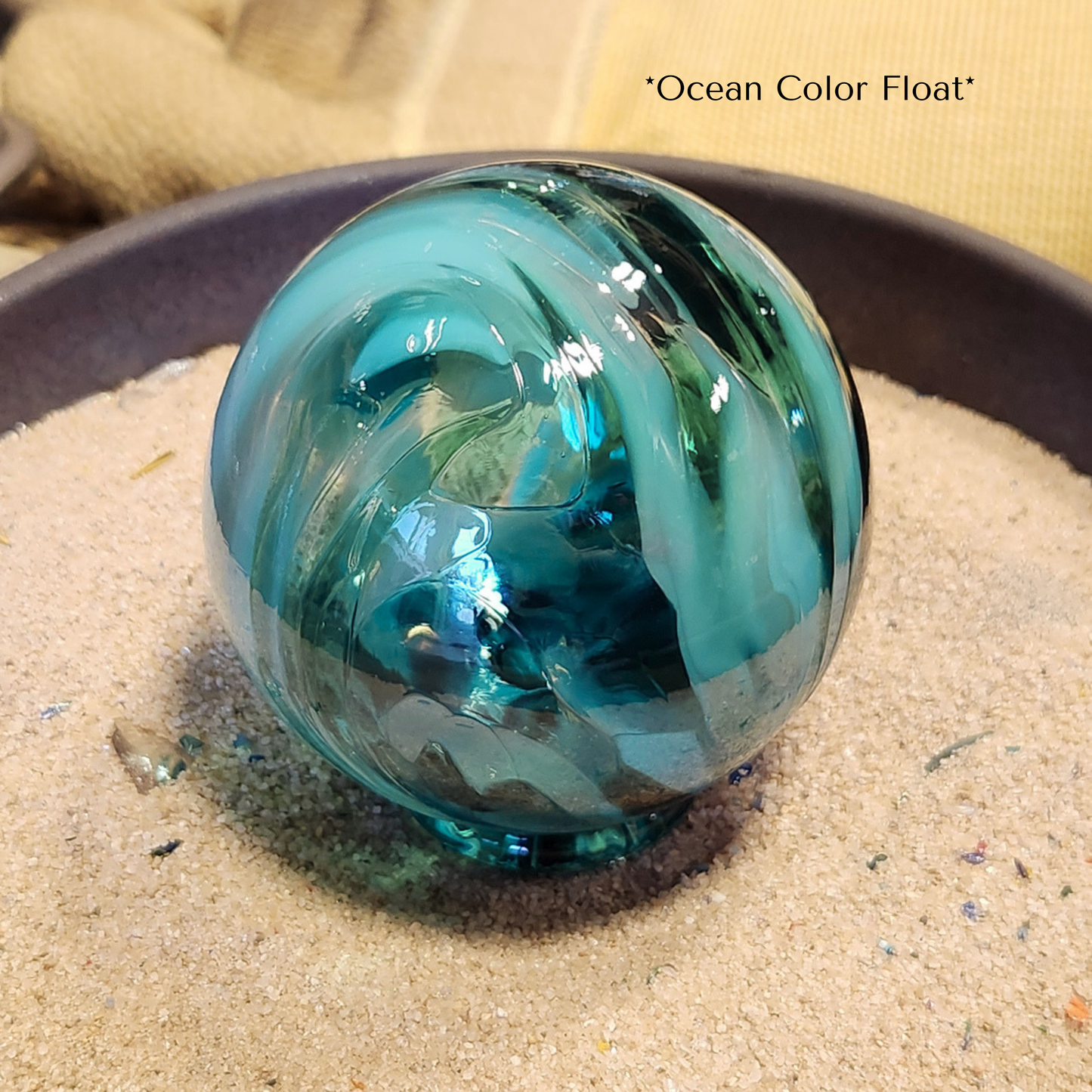 multiple shades of teal blown glass float sitting in a small dish of sand in the artists studio.